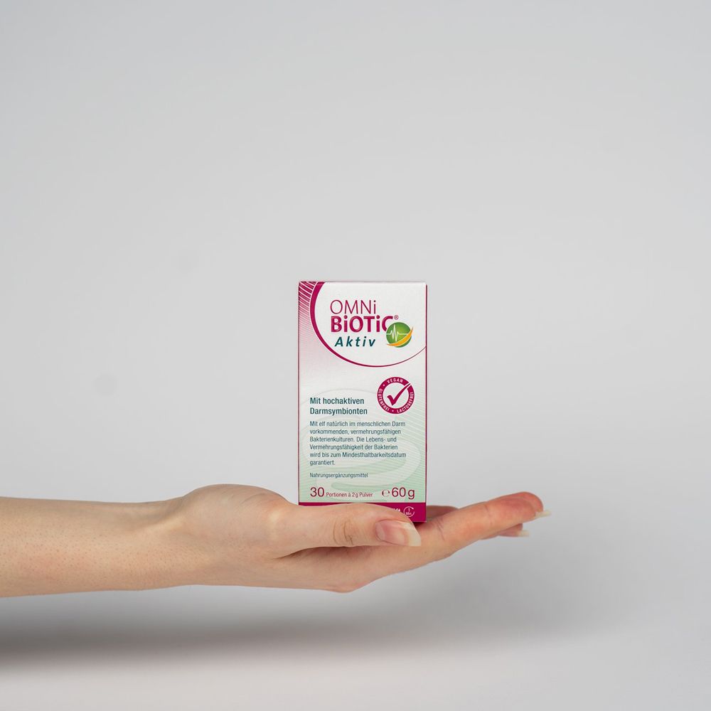 Hand hält eine OMNi BiOTiC Aktiv Kartonverpackung. 30 Portionen à 2g Pulver, 60g. Vegan, glutenfrei, laktosefrei.