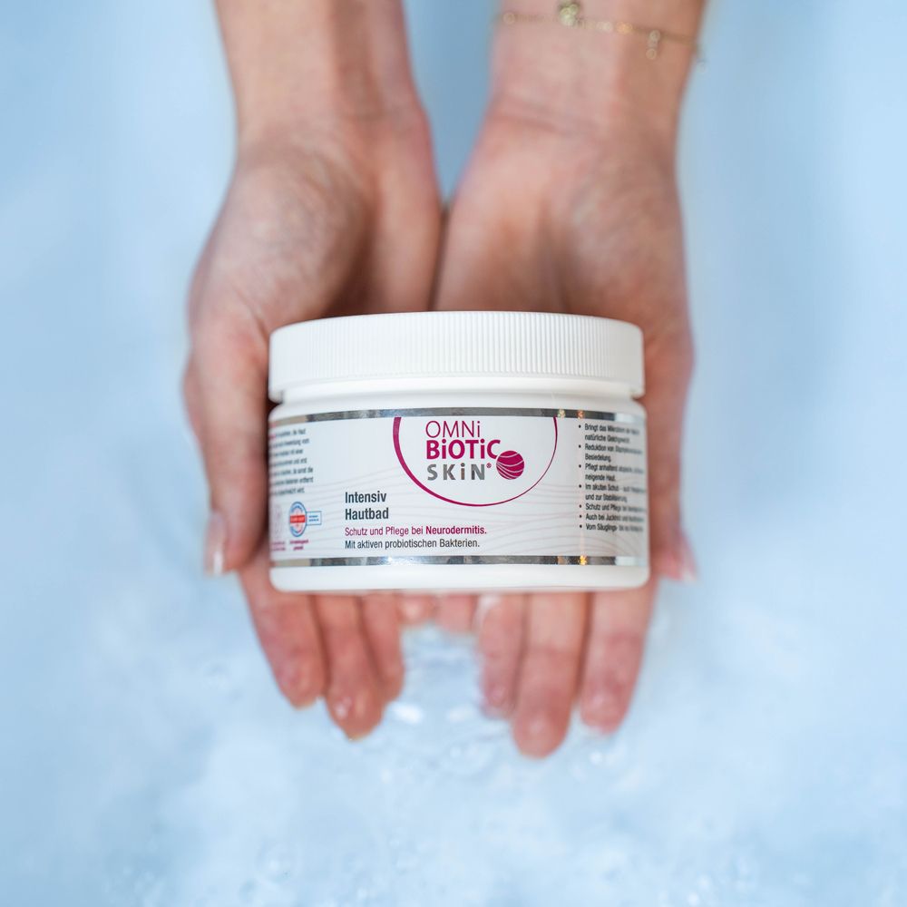 Weißes Gefäß mit OMNi-BiOTiC SKIN® Intensiv Hautbad, gehalten in Händen über blauem Wasser.