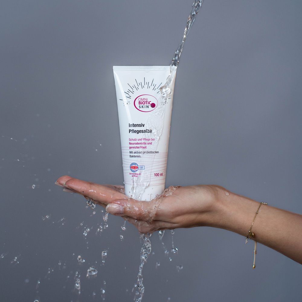 Weiße Tube OMNi-BiOTiC SKiN® Intensiv-Pflegesalbe, gehalten von einer Hand. Wasser fließt über die Tube.