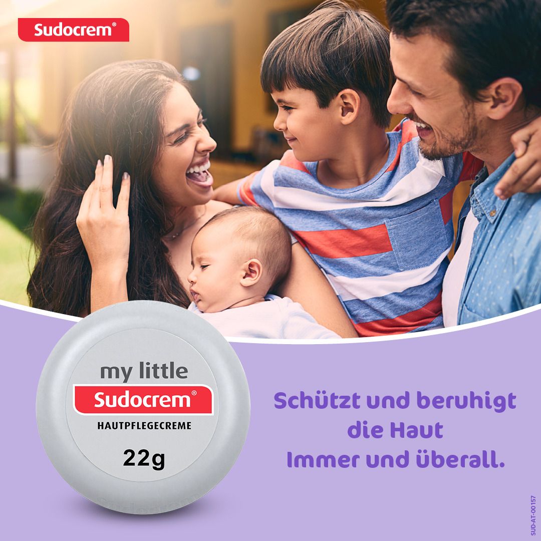 Runde, weiße Dose mit rotem und weißem Etikett. Aufschrift: my little Sudocrem, Hautpflegecreme, 22g. Familie im Hintergrund.