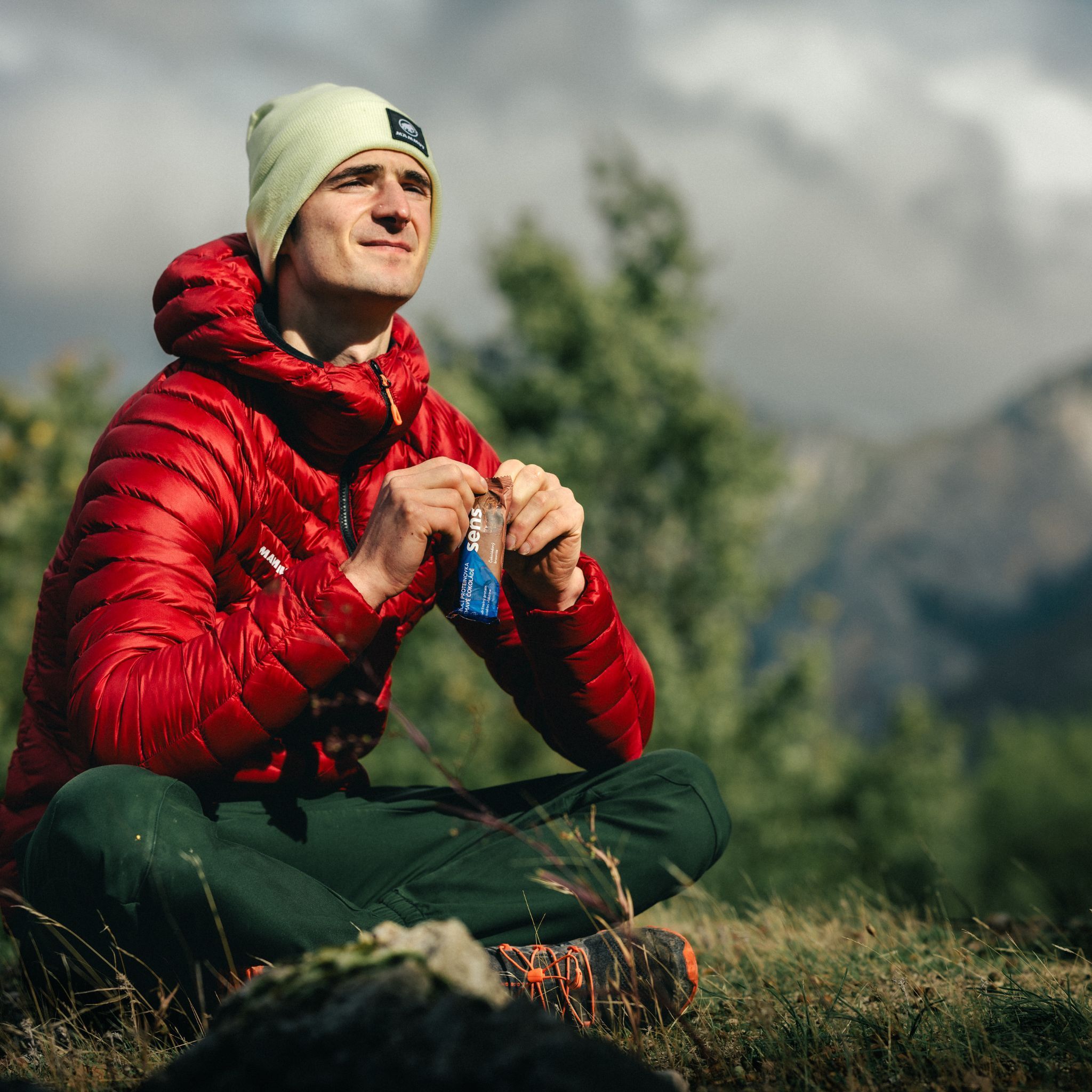 Mann sitzt im Freien und isst einen Proteinriegel. Er trägt eine rote Jacke und eine Mütze. Im Hintergrund Berge.