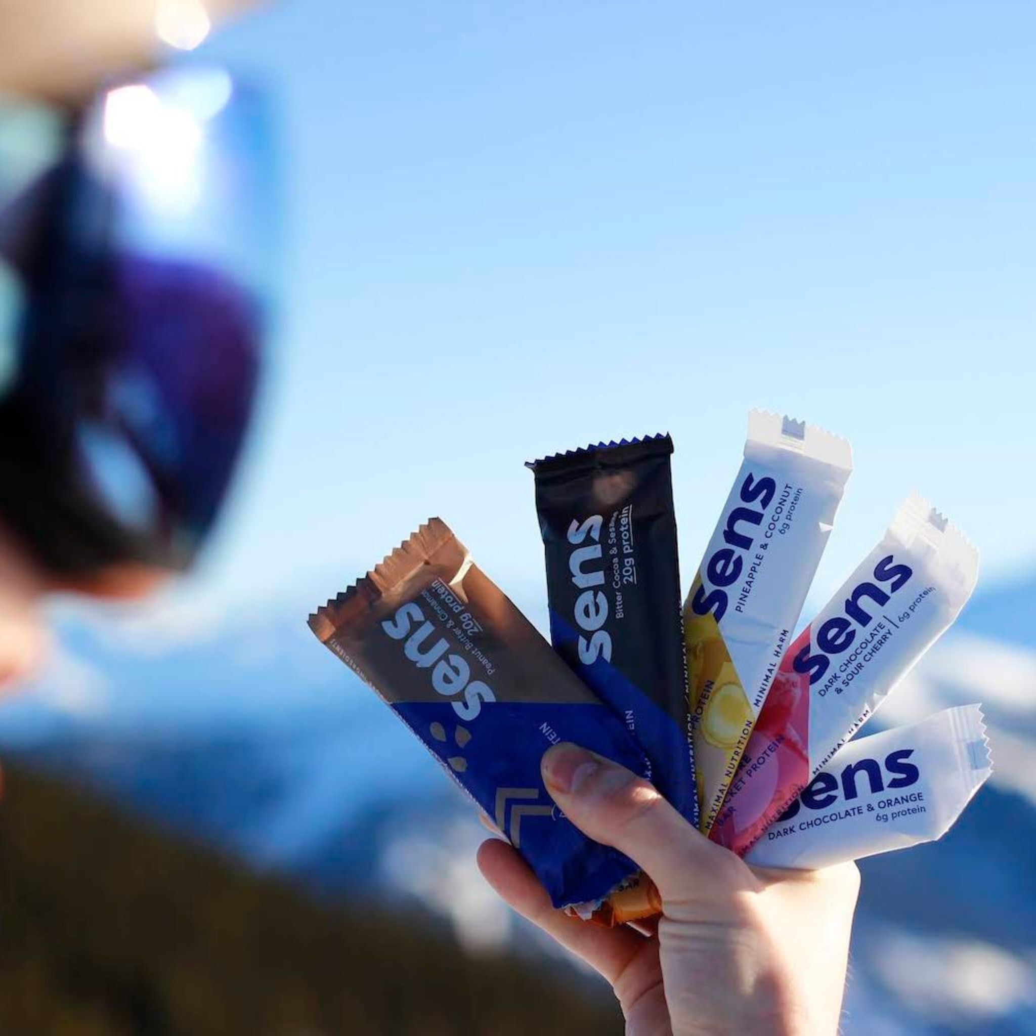 Hand hält mehrere Proteinriegel. Verschiedene Geschmacksrichtungen. Aufschrift: Sens. Hintergrund: verschwommene Berglandschaft.