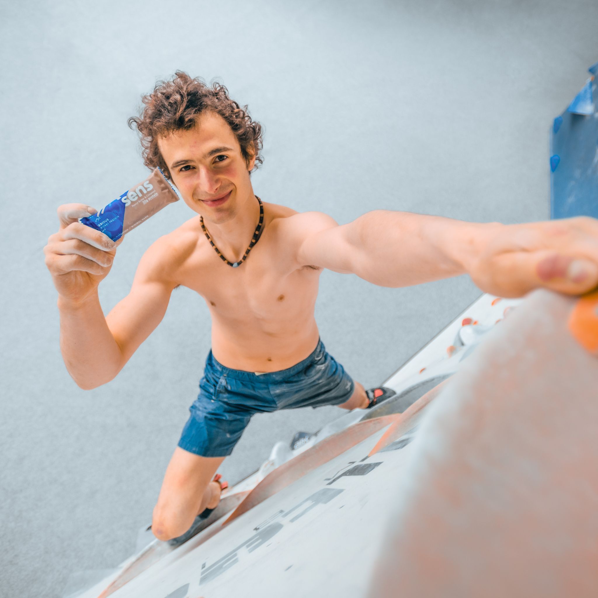 Mann hält eine Proteinriegel. Er ist barfuß und trägt Shorts. Im Hintergrund eine Kletterwand.