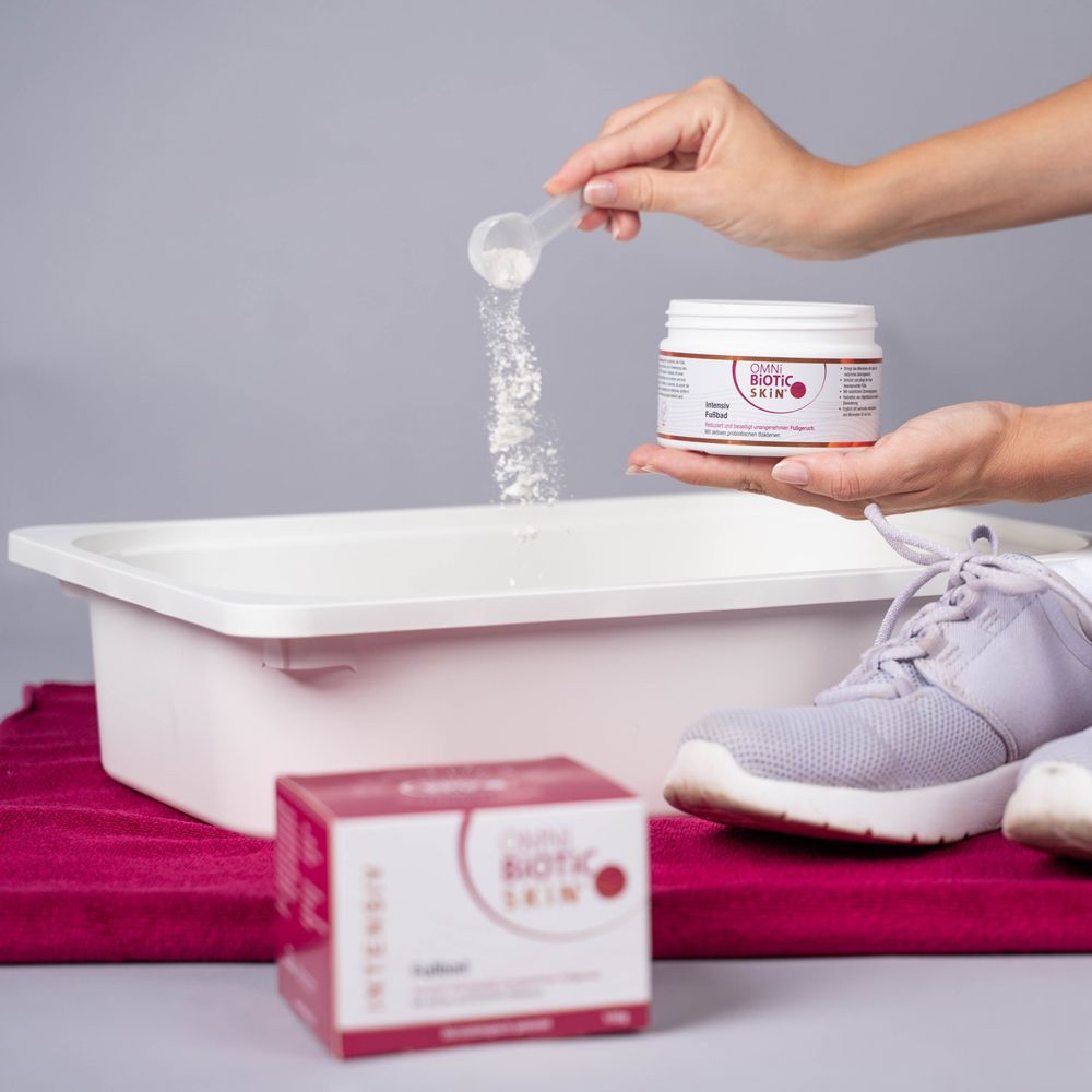 Person hält einen Messlöffel über eine Wanne. Daneben eine Dose und eine Schachtel OMNi-BiOTiC SKiN®. Schuhe liegen daneben.