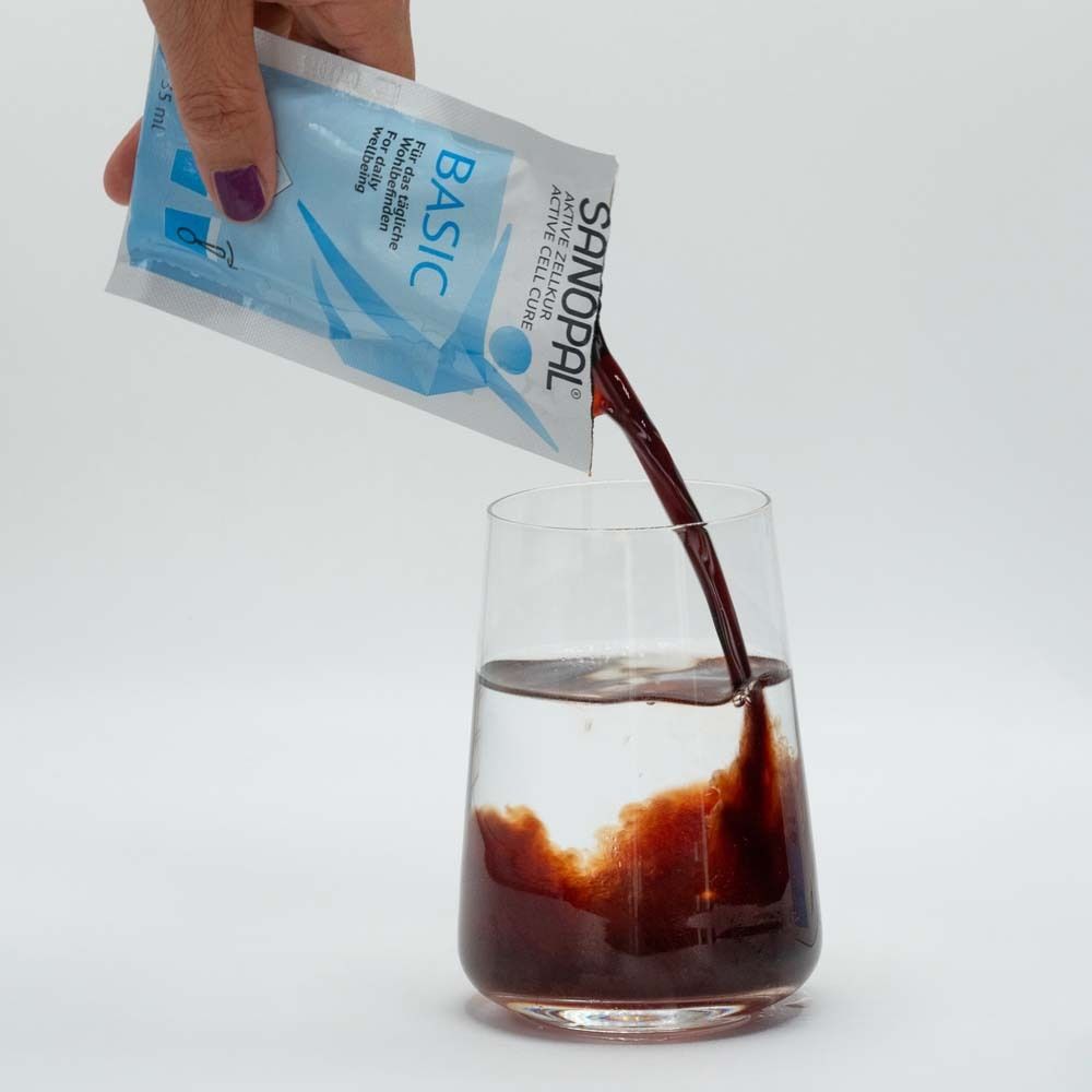 Eine Hand gießt eine braune Flüssigkeit aus einem Beutel in ein Glas Wasser. Aufschrift: SANOPAL BASIC.