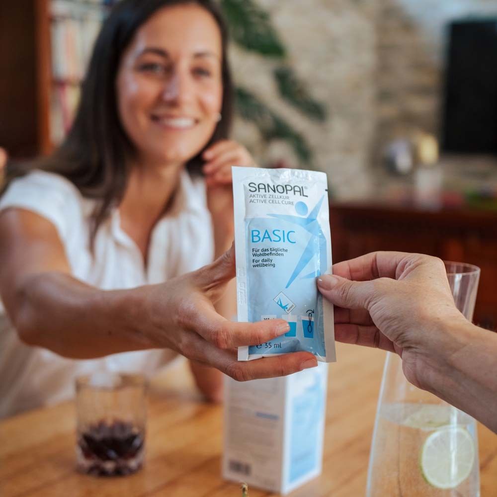 Zwei Hände reichen sich einen Beutel. Aufschrift: SANOPAL BASIC. Im Hintergrund ein Glas Wasser und eine Kanne.