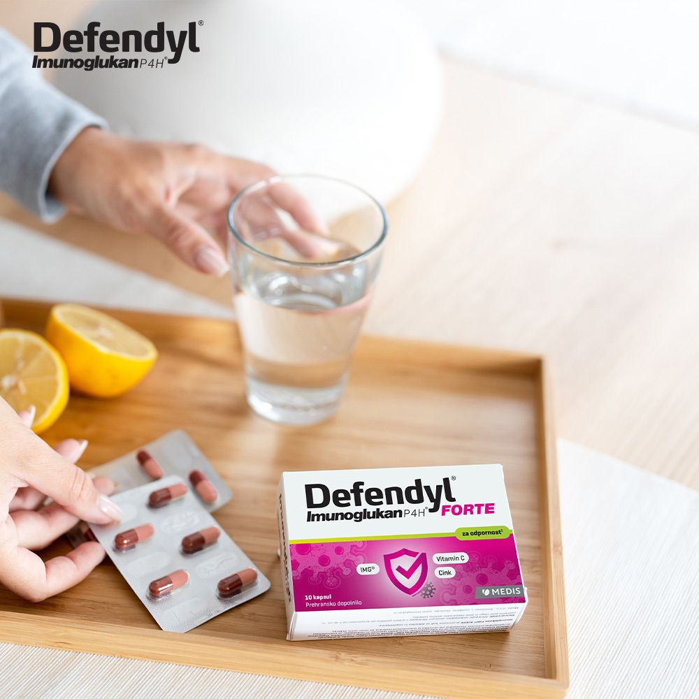 Hand nimmt Kapseln aus Blister. Defendyl-Imunoglukan P4H FORTE Packung und Glas Wasser auf Tablett.