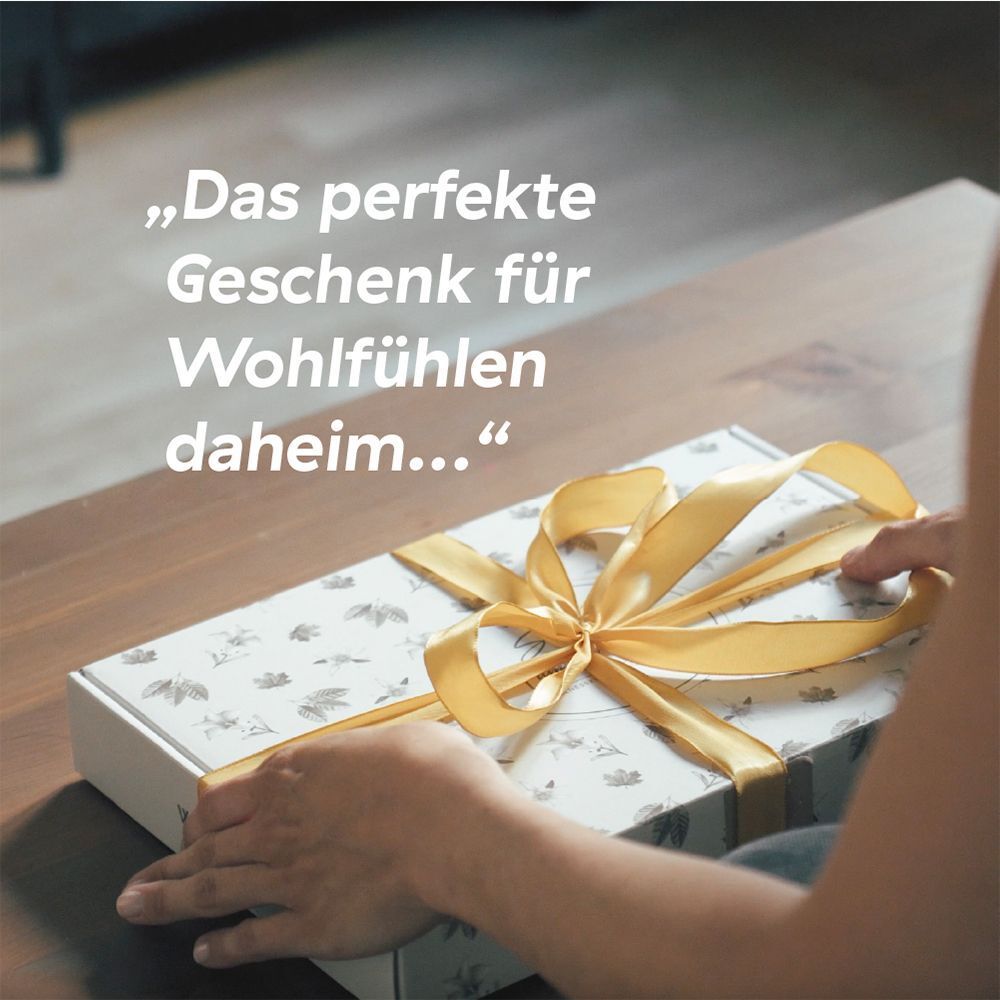 Person öffnet Geschenkbox mit Schleife. Text: Das perfekte Geschenk für Wohlfühlen daheim...