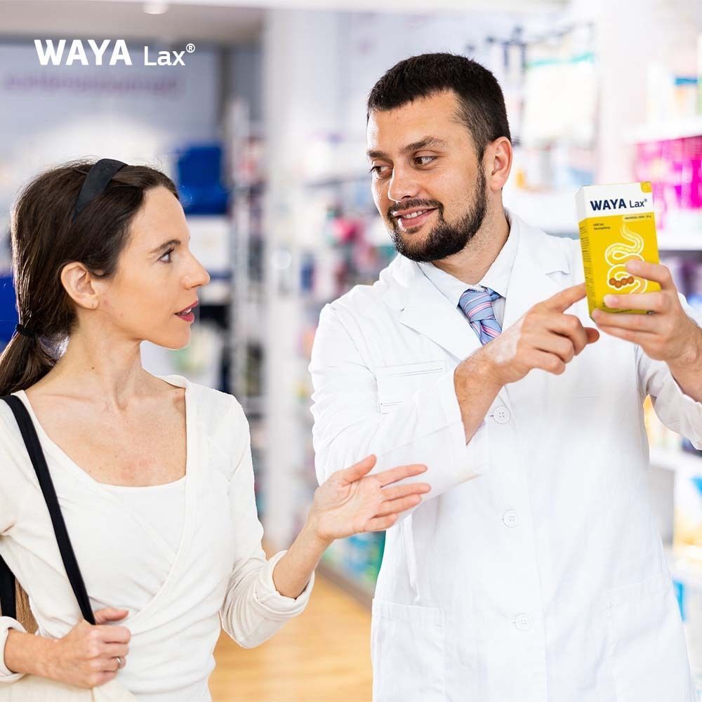 Apotheker zeigt WAYA Lax® Packung einer Kundin. Beide lächeln. Hintergrund: Apothekenregale.