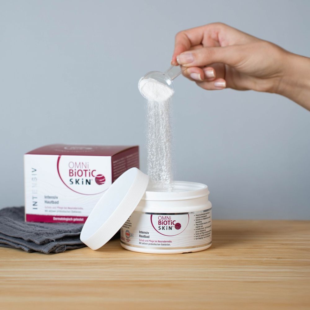 Eine Hand schüttet Pulver aus einem Messlöffel in ein offenes, weißes Gefäß. Daneben steht eine Produktverpackung.