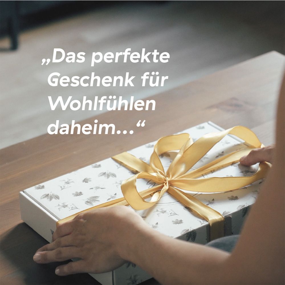 Hände öffnen eine Geschenkbox mit Schleife, Text: 'Das perfekte Geschenk für Wohlfühlen daheim...'