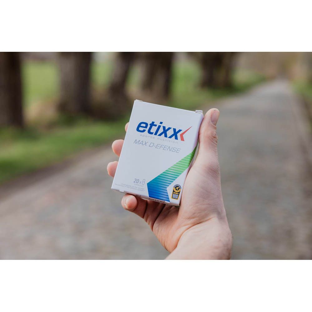 Hand hält eine kleine weiße Schachtel mit "etixx MAX D-EFENSE"-Logo. Hintergrund: verschwommener Weg im Freien.