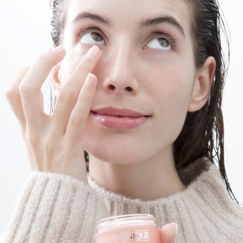 Frau hält eine rosa Creme-Dose von CLINIQUE. Sie berührt ihre Augenpartie. Hintergrund: hell.