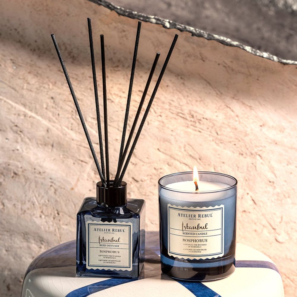 Duftkerze und Diffusor. Kerze mit Flamme, blaues Glas. Diffusor mit schwarzen Stäbchen, blaues Glas. Auf Tisch.
