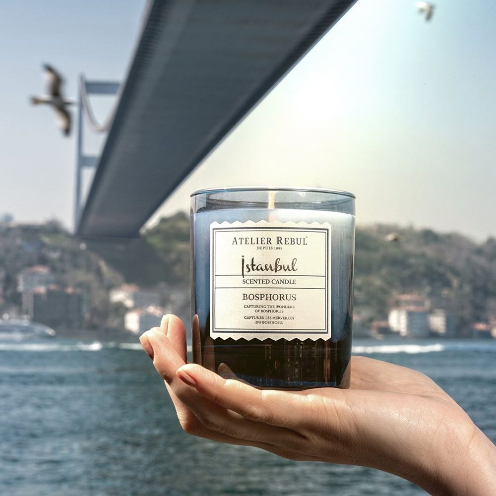 Hand hält Kerze. Kerze in blauem Glas, weißes Etikett. Hintergrund: Brücke, Meer, Himmel.