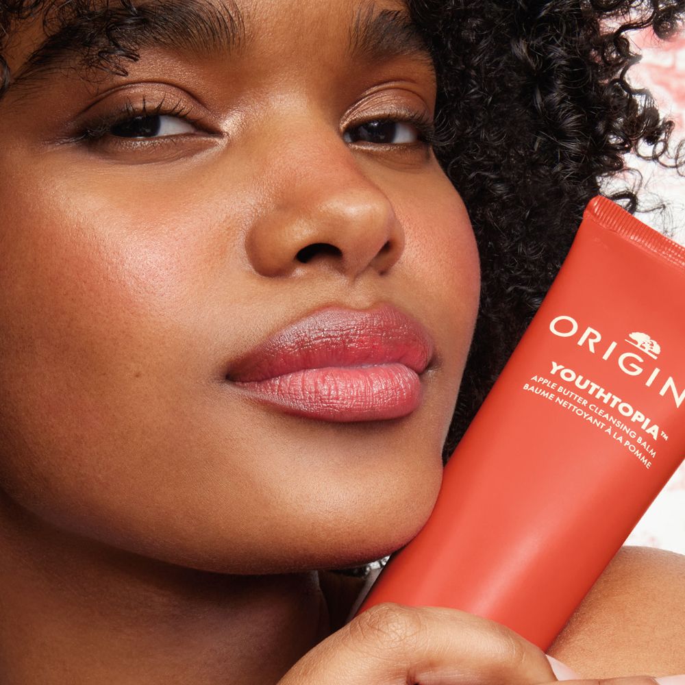 Frau hält eine rote Tube. Origins Youthtopia Apple Butter Cleansing Balm ist sichtbar.