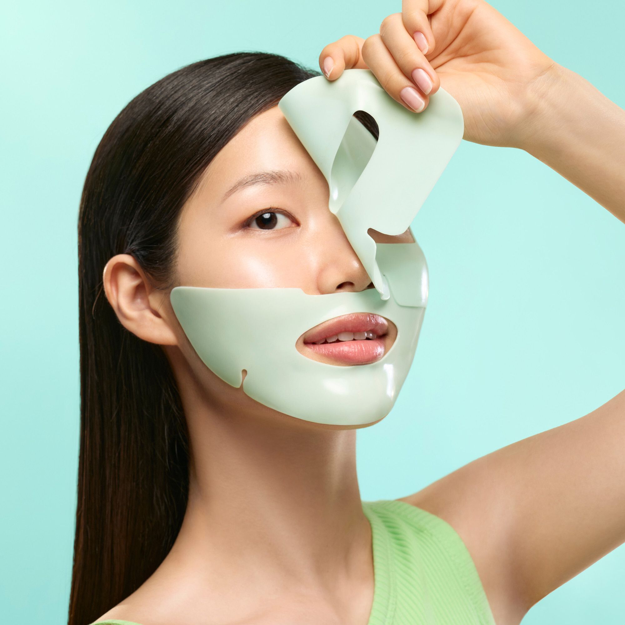 Junge Frau mit Maske. Maske in zwei Teilen, hellgrün. Frau hält Maske an. Grüner Hintergrund.