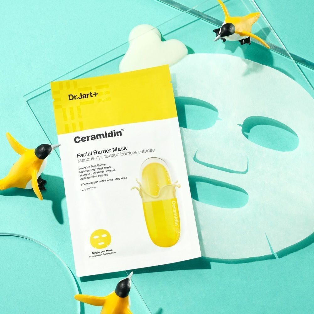 Gelbe Dr.Jart+ Ceramidin Gesichtsmaske auf blauem Hintergrund. Aufschrift: Facial Barrier Mask. Mit Dekoration.