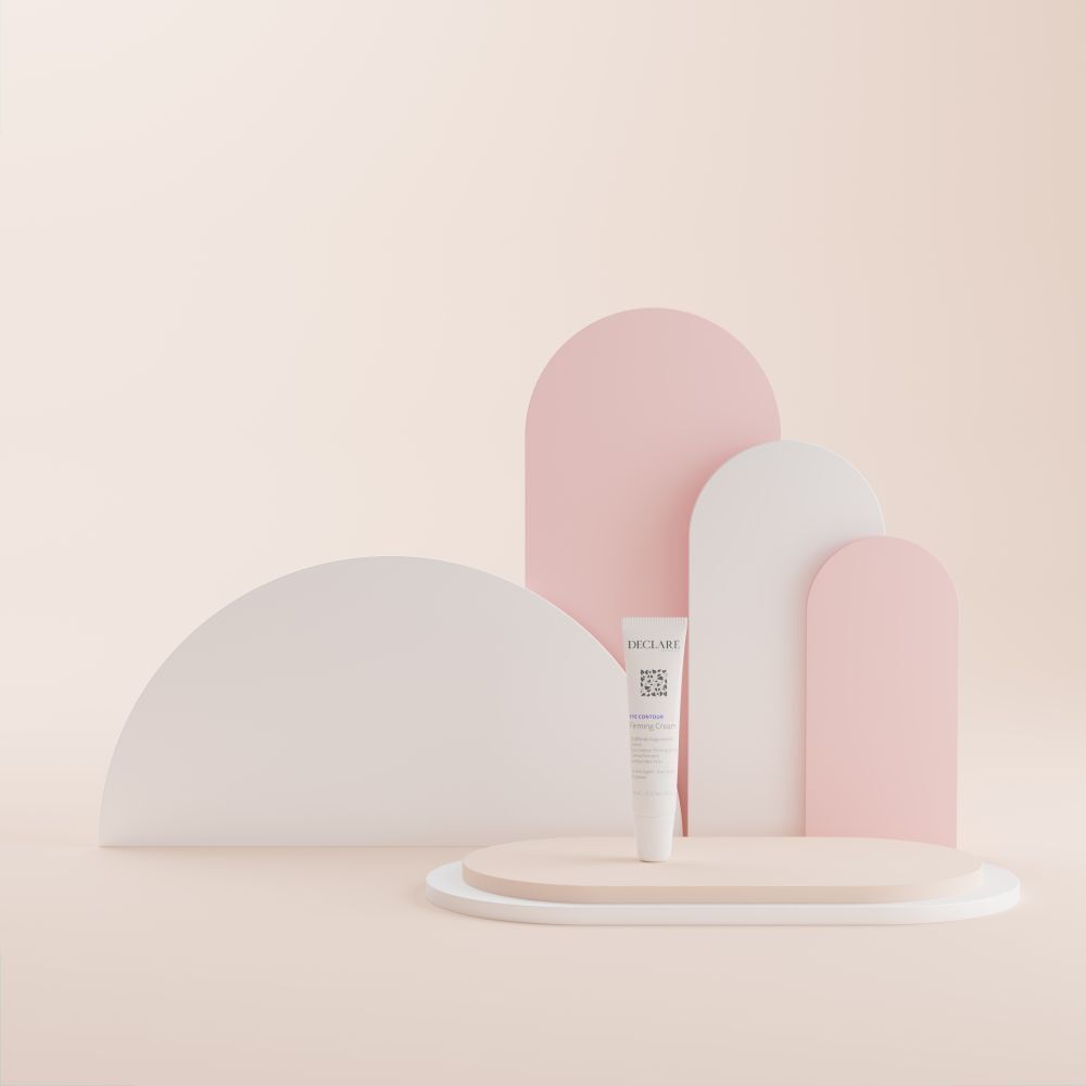 Weiße Tube DECLARÉ Eye Contour Firming Cream auf Podest. Hintergrund mit Formen in Rosa und Weiß.