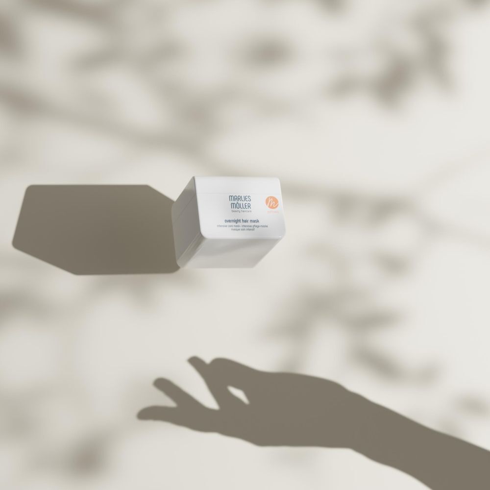 Weißer Behälter mit Marlies Möller Overnight Hair Mask, vor Schatten. Hand im Schatten.