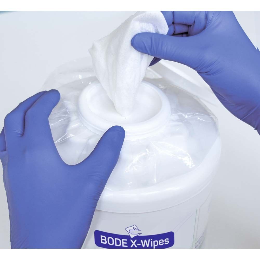 Weiße Tücher werden aus einem weißen Behälter entnommen. Hände mit blauen Handschuhen. Auf dem Behälter steht BODE X-Wipes.