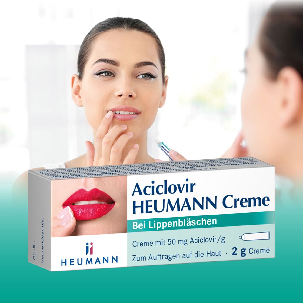 Frau trägt Creme auf die Lippen auf. Im Hintergrund: Schachtel Aciclovir Heumann Creme. Bei Lippenbläschen.