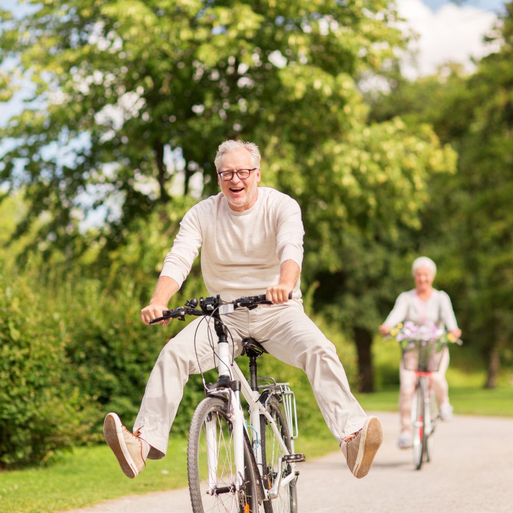 Älterer Mann auf einem Fahrrad, springend. Eine Frau fährt im Hintergrund Fahrrad.