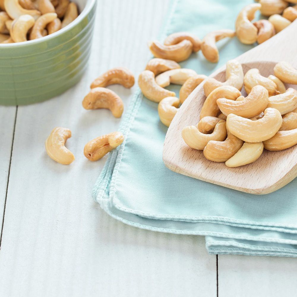Cashewnüsse auf einem Holzlöffel und auf einem blauen Tuch. Grüne Schale im Hintergrund.