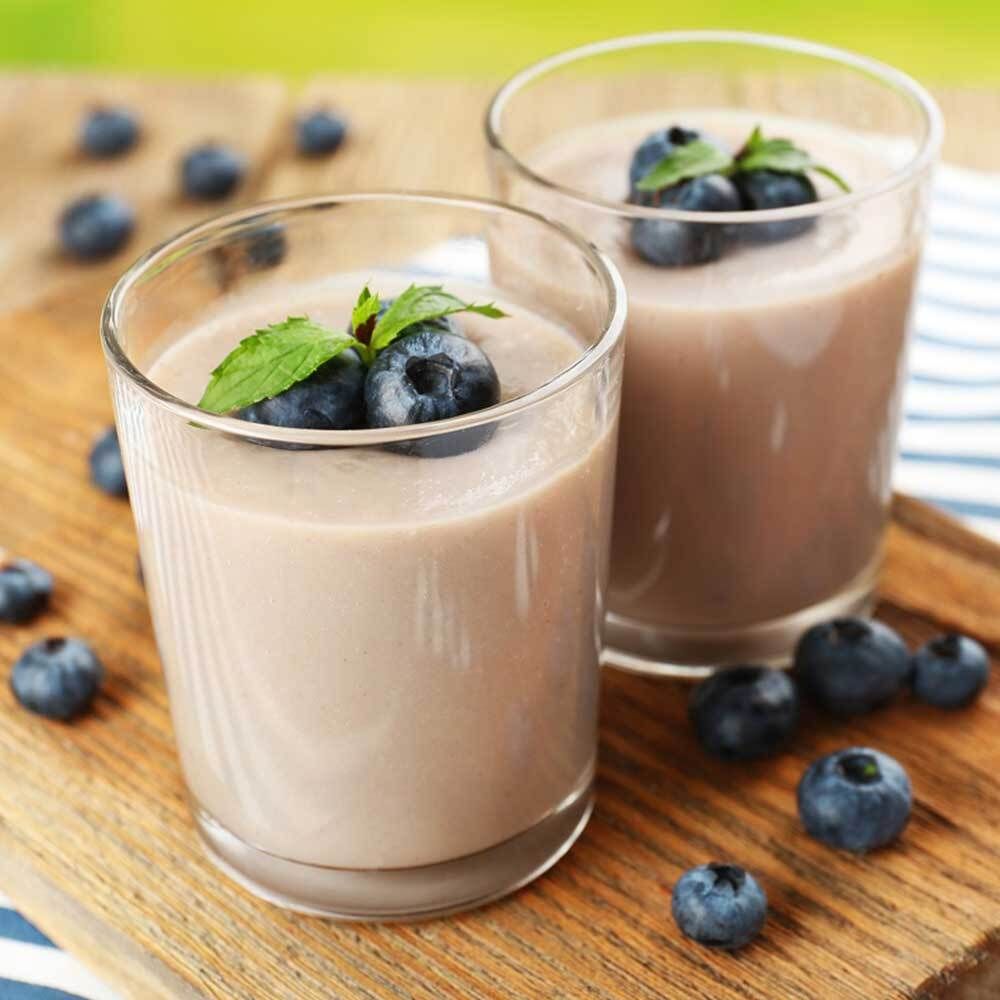 Zwei Gläser mit lila Shake, Blaubeeren und Minze auf Holzbrett. Blaubeeren verstreut.