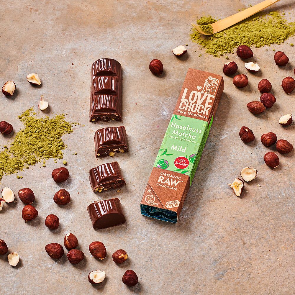 Schokoladenriegel und -stücke, Haselnüsse und Matcha-Pulver auf Untergrund. Verpackung: LOVECHOCK, Haselnuss Matcha Mild, Organic Raw Chocolate.