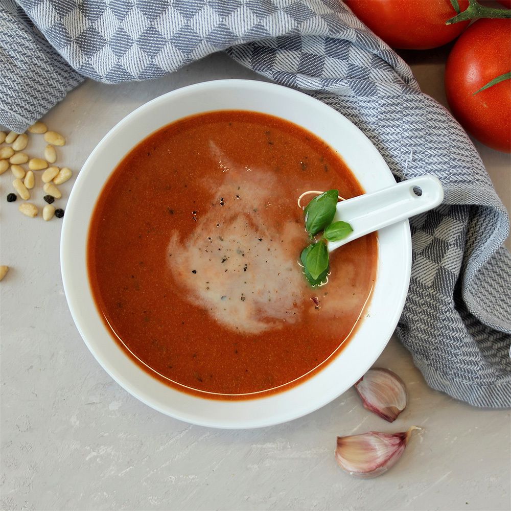 Tomatensuppe in weißer Schale mit Löffel. Garniert mit Basilikum. Auf grauem Untergrund mit Tomaten, Knoblauch und Pinienkernen.