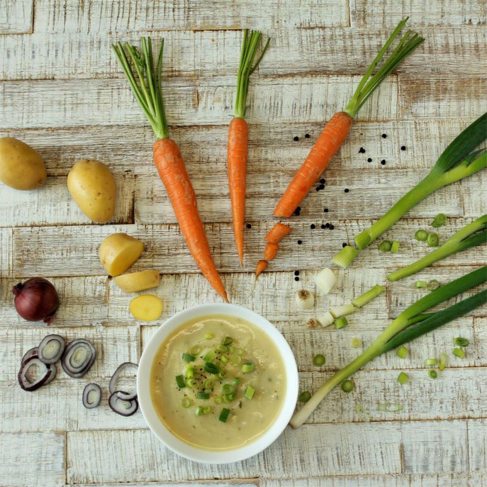 Schüssel Kartoffelsuppe mit Frühlingszwiebeln. Daneben Karotten, Kartoffeln, Zwiebeln, Pfefferkörner und Lauch auf Holzbrett.