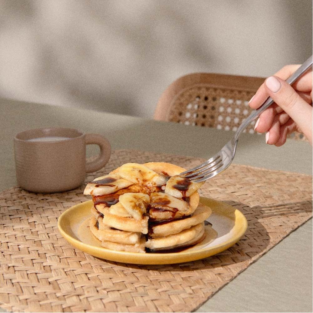 Pfannkuchen mit Bananenscheiben auf einem Teller. Eine Hand hält eine Gabel. Im Hintergrund eine Tasse.