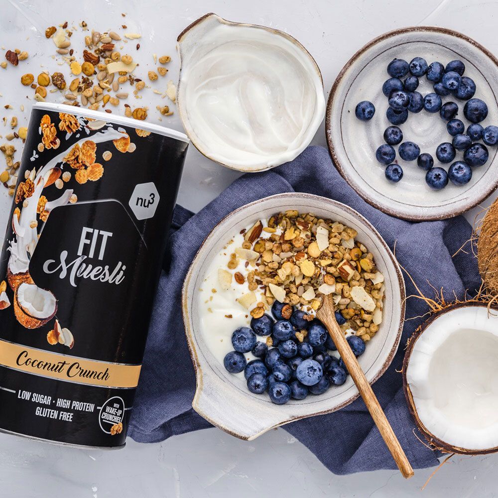 Schwarze Dose nu3 Fit Müsli, Coconut Crunch, neben Schüssel mit Müsli, Blaubeeren, Joghurt. Kokosnuss und Holzlöffel auf Tisch.