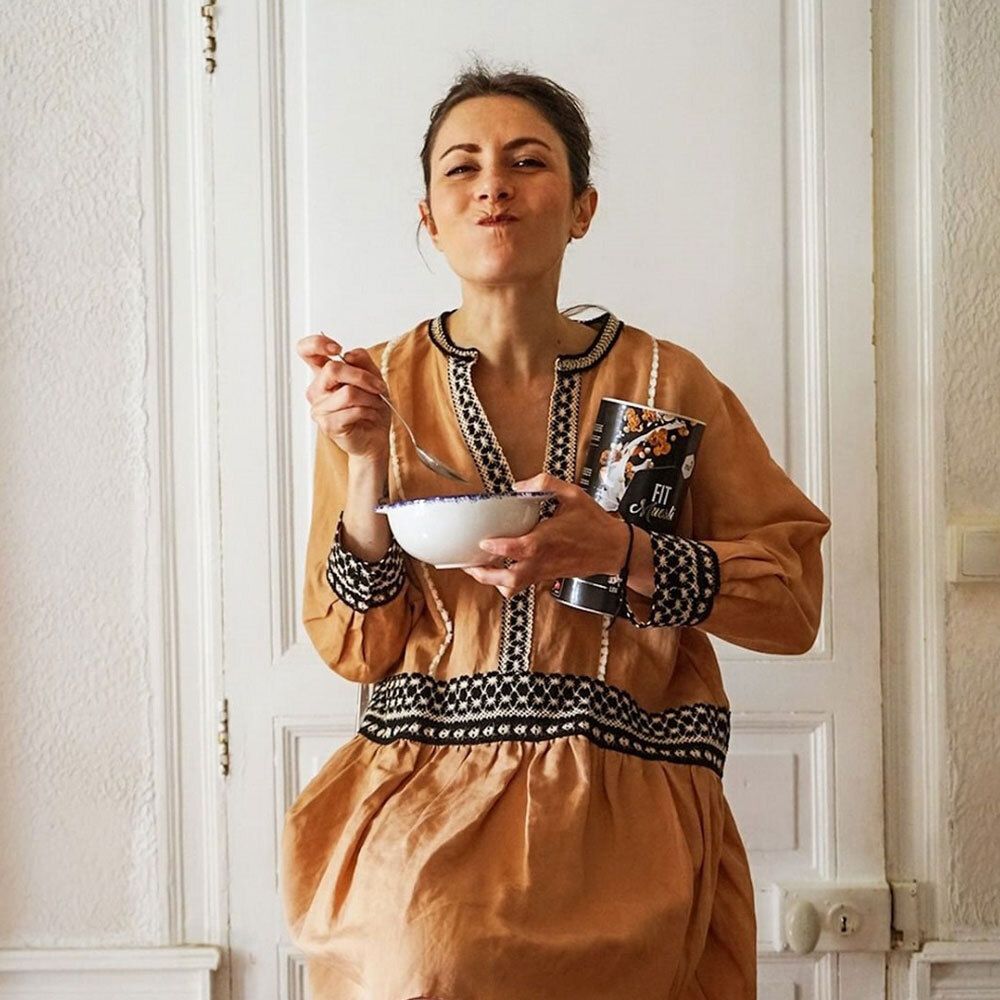 Frau mit beigem Kleid hält Dose nu3 Fit Müsli, Coconut Crunch und isst aus Schüssel. Tür im Hintergrund.
