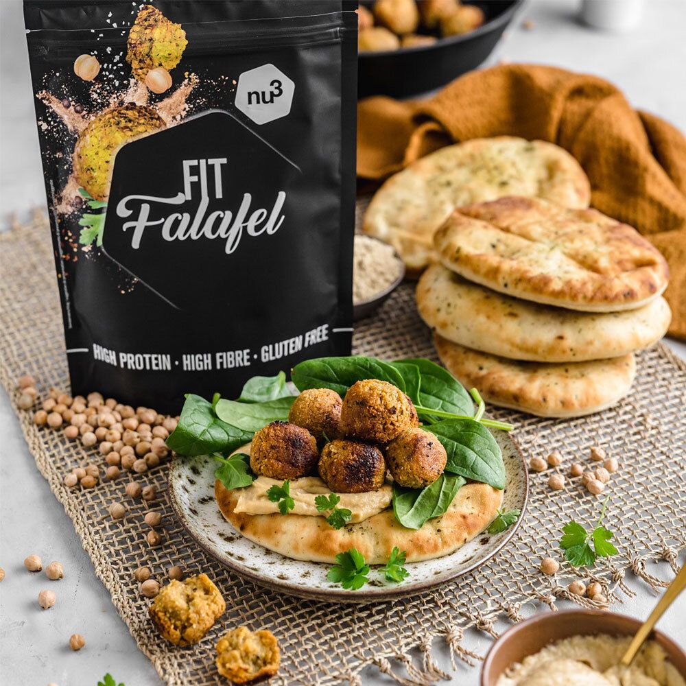 nu3 Fit Falafel-Packung neben einem Teller mit Falafel, Hummus und Brot. Kichererbsen und Brotstücke liegen daneben.