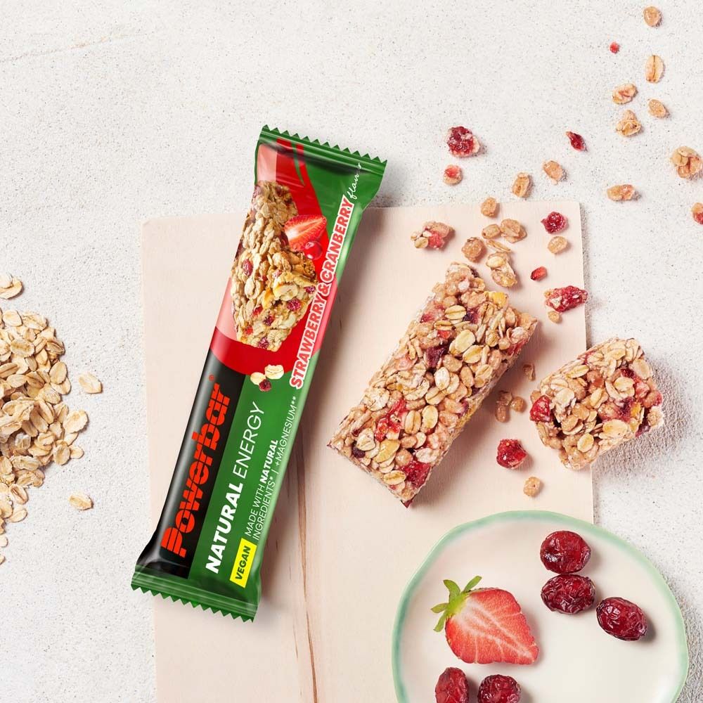PowerBar Natural Energy Riegel, Erdbeer-Cranberry. Auf hellem Untergrund. Cranberrys, Erdbeere und Müsli-Stücke daneben.