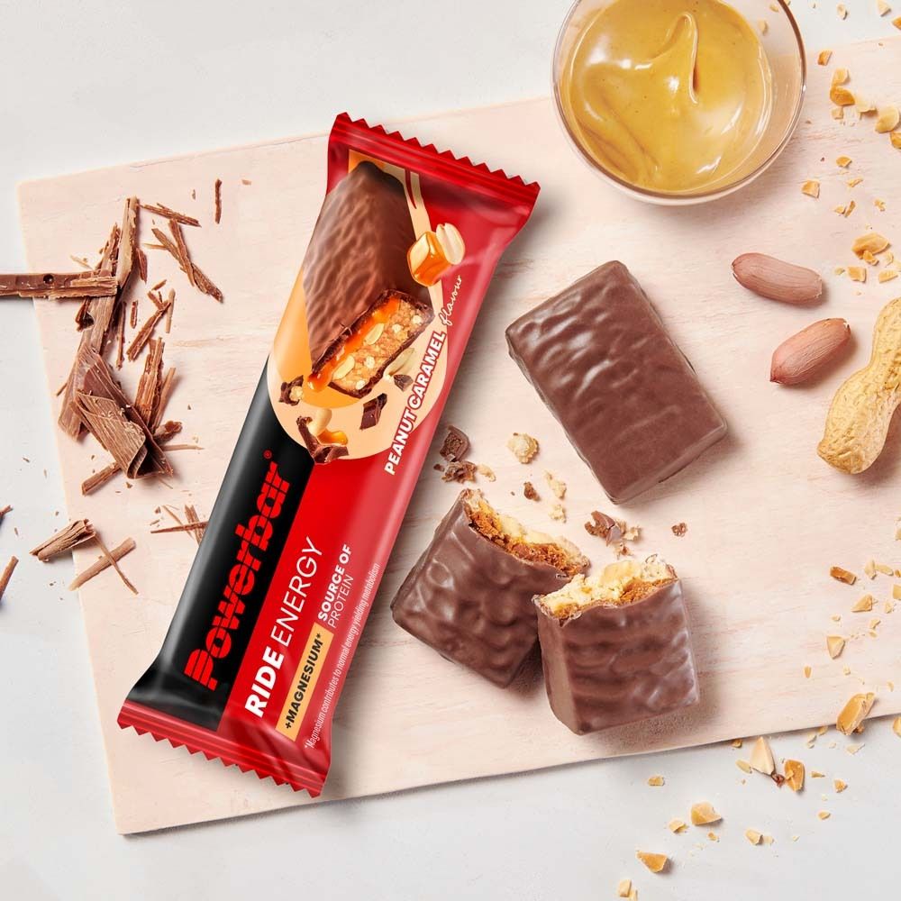 PowerBar Ride Energy Riegel, Peanut Caramel. Auf Holzbrett. Geöffneter Riegel, Erdnüsse und Erdnussbutter im Hintergrund.