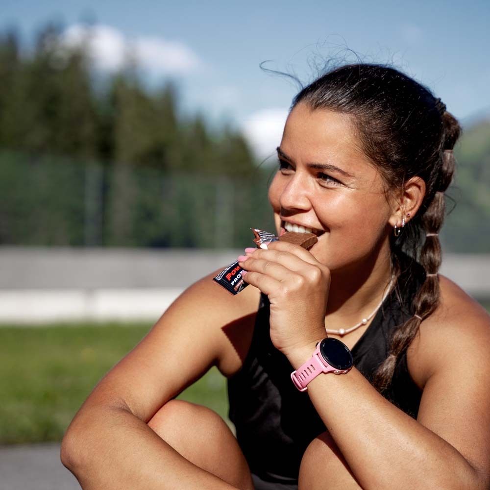 Frau isst Powerbar Protein+ Riegel. Sie sitzt draußen. Sie trägt eine Smartwatch.