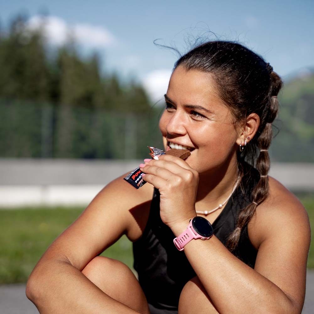 Frau isst Powerbar Protein+ Fibre Riegel. Rosa Verpackung. Sie trägt eine Smartwatch. Draußen, sonniger Tag.