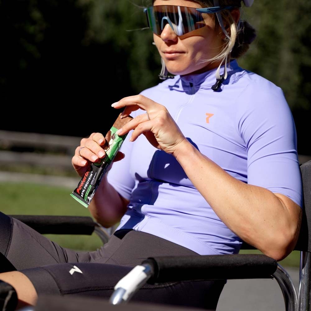 Person hält eine PowerBar 52% Protein Bar. Sie sitzt in einem Rollstuhl und trägt Sportkleidung und Sonnenbrille.