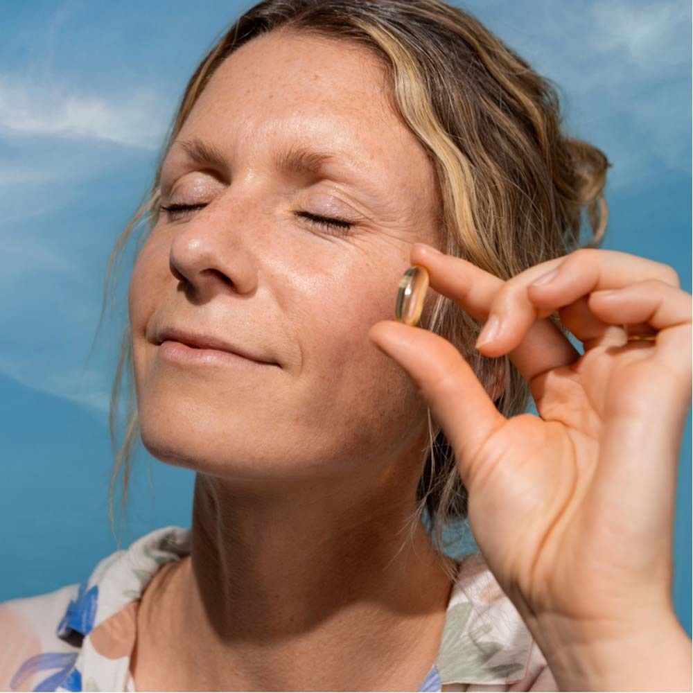 Frau hält eine goldfarbene Omega-3 Kapsel zwischen den Fingern. Blauer Himmel im Hintergrund.