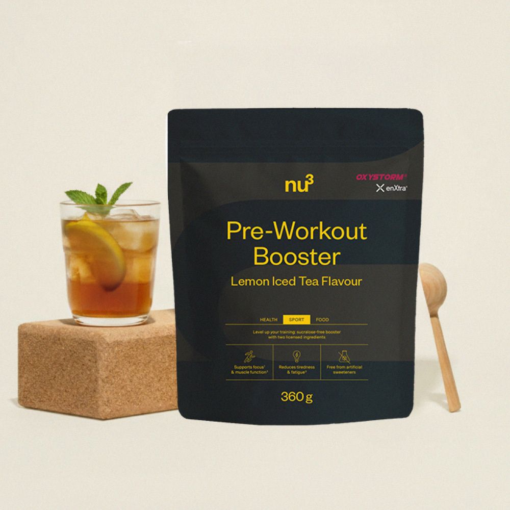 Schwarze Verpackung "nu3 Pre-Workout Booster" neben Glas mit Getränk, Holzlöffel. Aufschrift: Zitrone-Eistee-Geschmack.