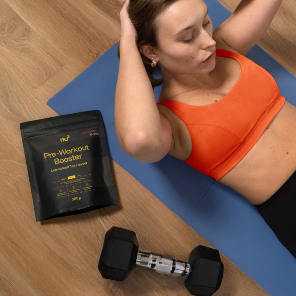 Frau trainiert auf einer blauen Matte. Neben ihr: "nu3 Pre-Workout Booster" und Hantel. Sport-Kontext.