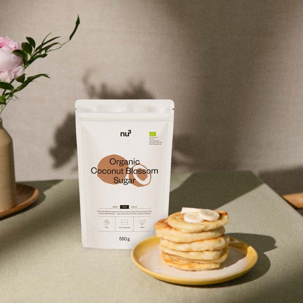 Produktverpackung neben Pfannkuchen auf gelbem Teller. Blüten in Vase. nu3 Organic Coconut Blossom Sugar, 550g.