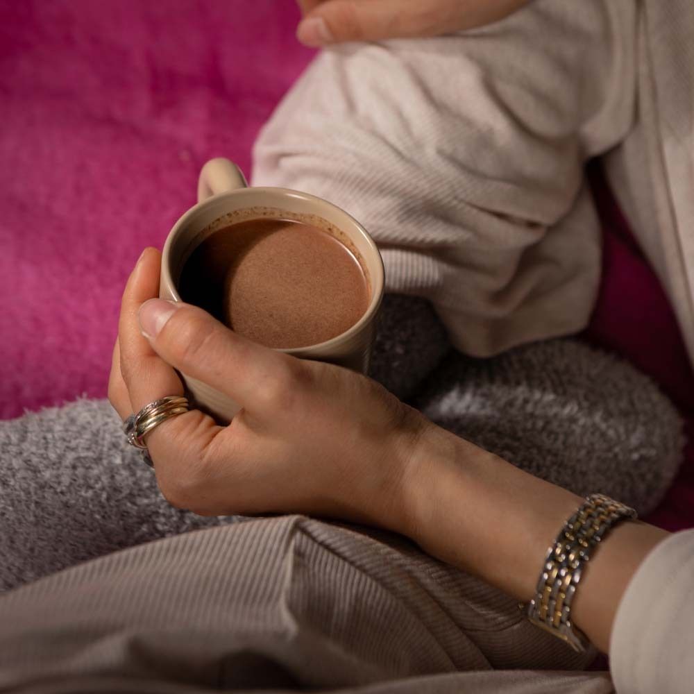 Hände halten eine Tasse Kakao. Person trägt Ringe und Armband. Hintergrund: rosa Decke.