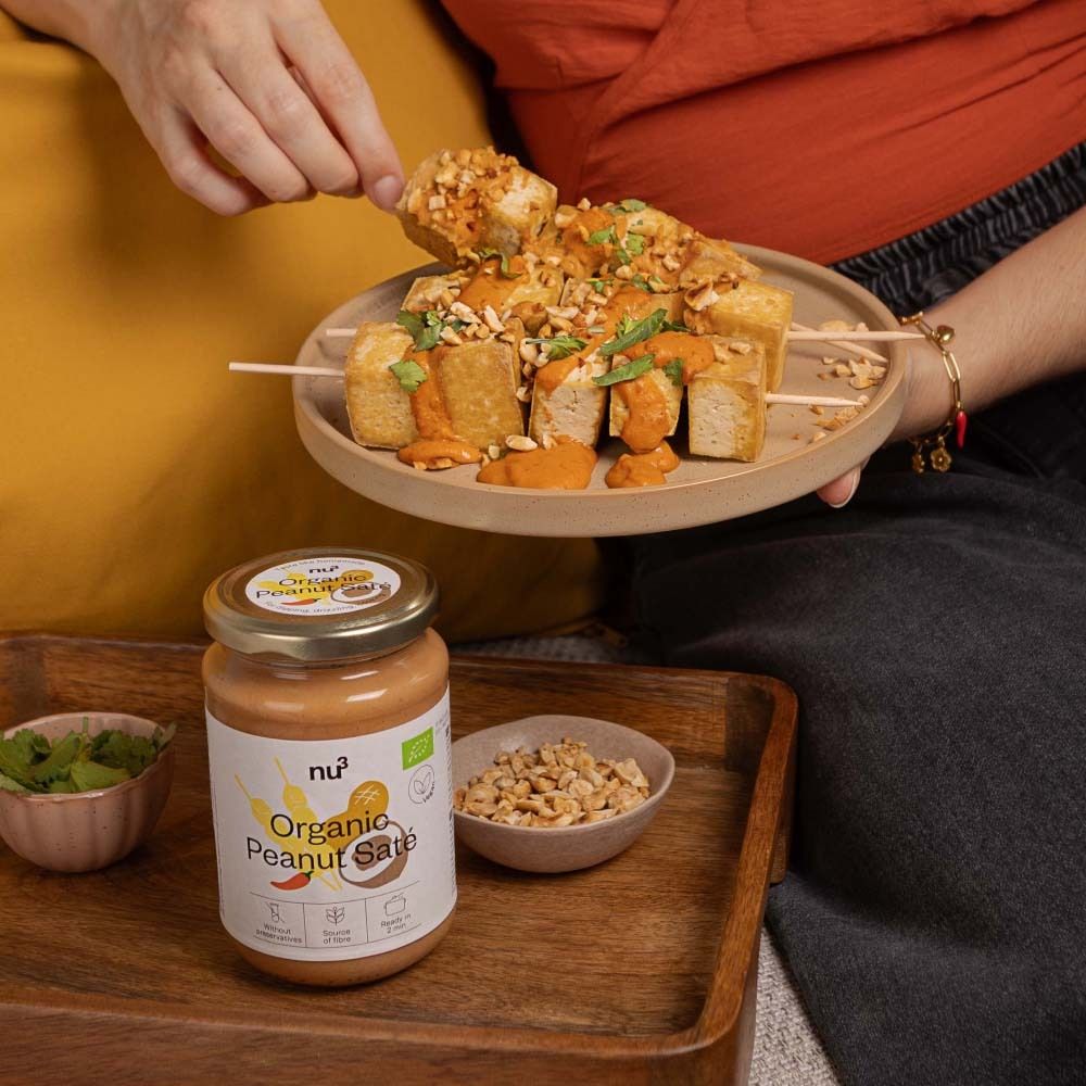 Person hält Spieß mit Tofu und Erdnuss-Saté-Sauce. Daneben Glas mit Sauce und Schale mit Erdnüssen. Aufschrift: Organic Peanut Saté.