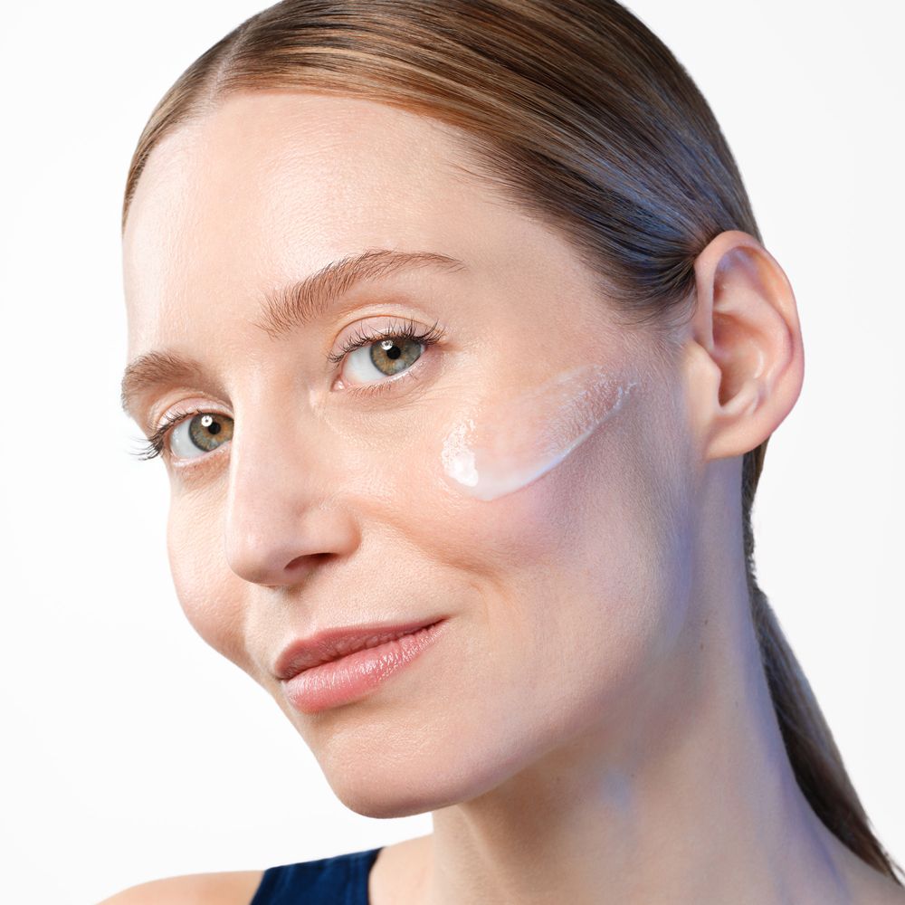 Frau mit Creme auf der Wange. Helle Haut, braune Haare. Blick in die Kamera.
