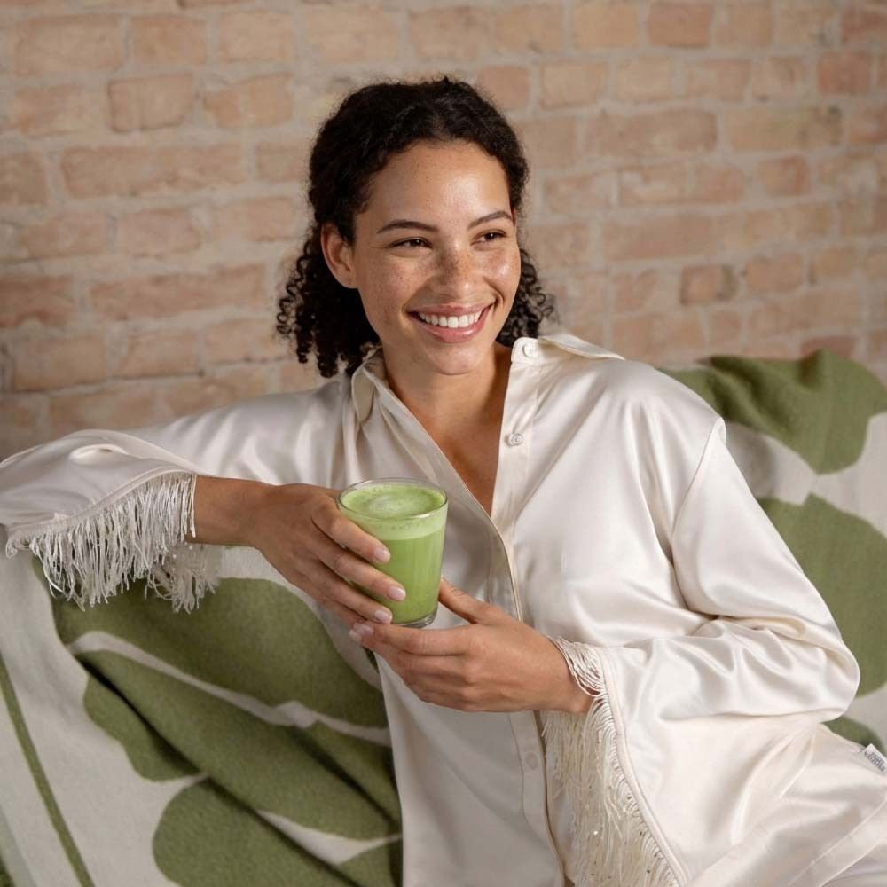 Frau sitzt auf Sofa, hält Glas Matcha Latte. Sie lächelt. Helle Kleidung, Decke mit grünem Muster.