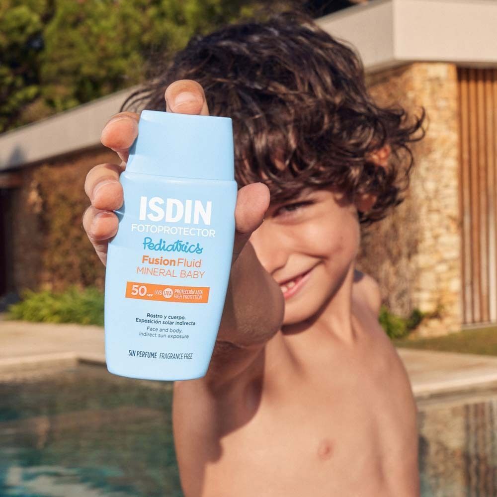 Ein Kind hält eine blaue Flasche mit weißem Deckel. Aufschrift: ISDIN Fotoprotector Pediatrics Fusion Fluid Mineral Baby LSF 50.